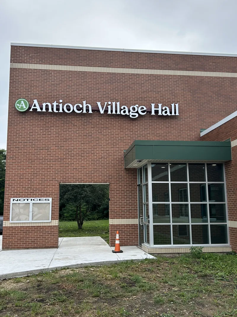 Antioch Village Hall exterior — dimensional white lettering and a green circular 'A' logo mounted on a red brick facade above a glass entry.