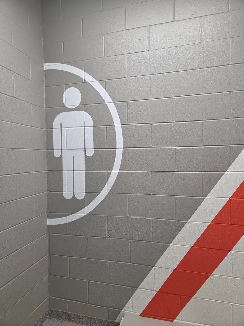 School athletic facility stairwell — large painted runner/swift figure inside a half-arc emblem on a soft-gray block wall, with a bold red diagonal stripe accent overhead.