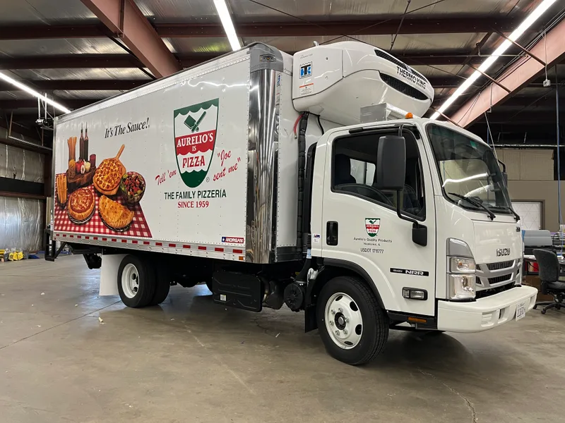 Aurelio's Pizza refrigerated box truck — modern white Isuzu NRR with a full-side wrap featuring the Aurelio's shield logo, 'It's The Sauce!' tagline, photorealistic pizza spread on a red-and-white checkered tablecloth, and 'THE FAMILY PIZZERIA SINCE 1959', photographed after install.