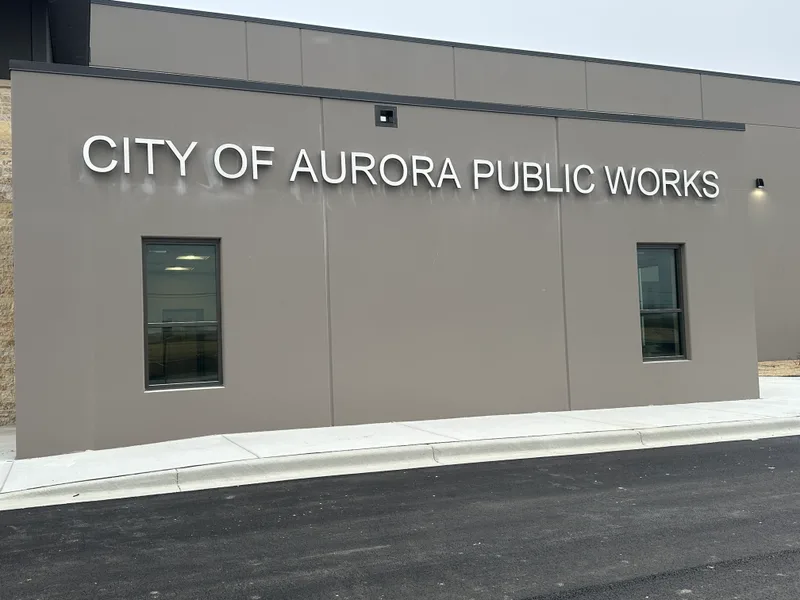 City of Aurora Public Works facility — clean large white dimensional letters reading 'CITY OF AURORA PUBLIC WORKS' mounted on the tan-stucco institutional building facade above a freshly-paved drive.