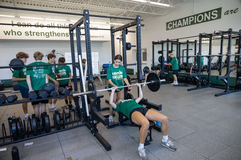 Boylan Catholic High School weight room — students using the bench rack with 'CHAMPIONS are...' and 'I can do all things through Christ who strengthens me — Philippians 4:13' wall graphics installed across the back wall.