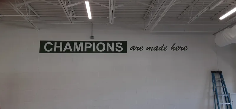 "CHAMPIONS are made here" — green block and script lettering applied high on the white wall of Boylan's new performance center.