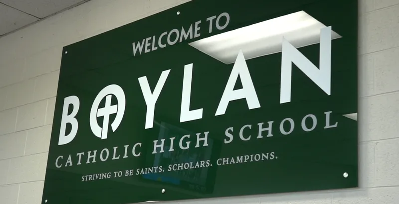 Boylan Catholic High School welcome sign — dimensional metallic letters reading 'WELCOME TO BOYLAN CATHOLIC HIGH SCHOOL — Striving to be Saints, Scholars, Champions' framed by a dark green panel with leaf accents.