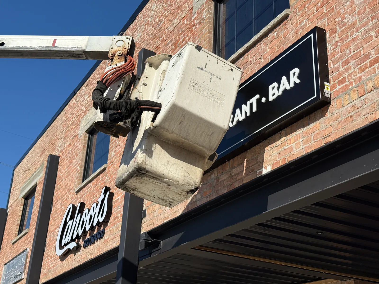 CorPro install-in-progress on Cahoots Rockford — Altec bucket-truck lift with an installer in the bucket mounting the dimensional 'Cahoots' script letters onto the historic brick facade.