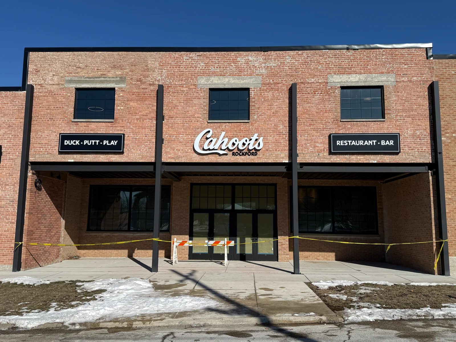 Cahoots Rockford storefront — freshly restored historic brick facade with dimensional white 'Cahoots' script logo (with 'ROCKFORD' beneath) flanked by black 'DUCK · PUTT · PLAY' and 'RESTAURANT · BAR' cabinet signs mounted above the entrance colonnade.