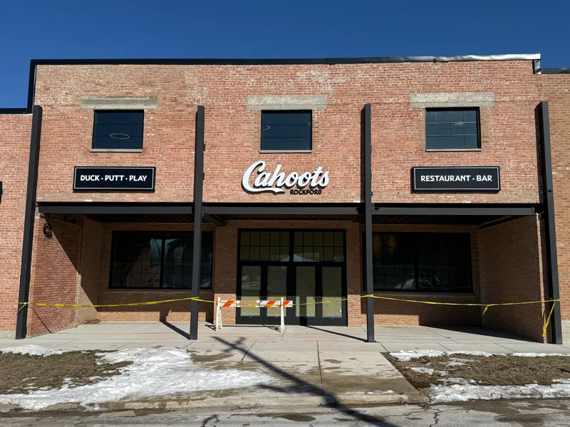 Cahoots Rockford storefront — freshly restored historic brick facade with dimensional white 'Cahoots' script logo (with 'ROCKFORD' beneath) flanked by black 'DUCK · PUTT · PLAY' and 'RESTAURANT · BAR' cabinet signs mounted above the entrance colonnade.