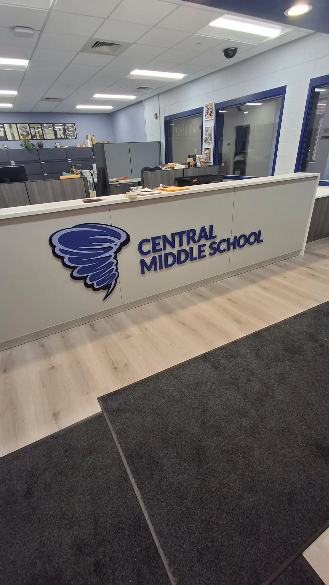 Central Middle School reception — dimensional blue tornado mascot logo + 'CENTRAL MIDDLE SCHOOL' block wordmark mounted on a light-gray reception counter, wood-look flooring in the foreground, staff office and school-spirit letters visible behind.