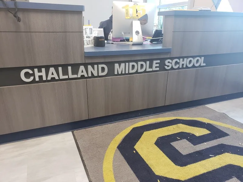 Challand Middle School reception desk — dimensional brushed-metal "CHALLAND MIDDLE SCHOOL" letters mounted to a wood-paneled front counter, with a branded school-mascot floor mat.
