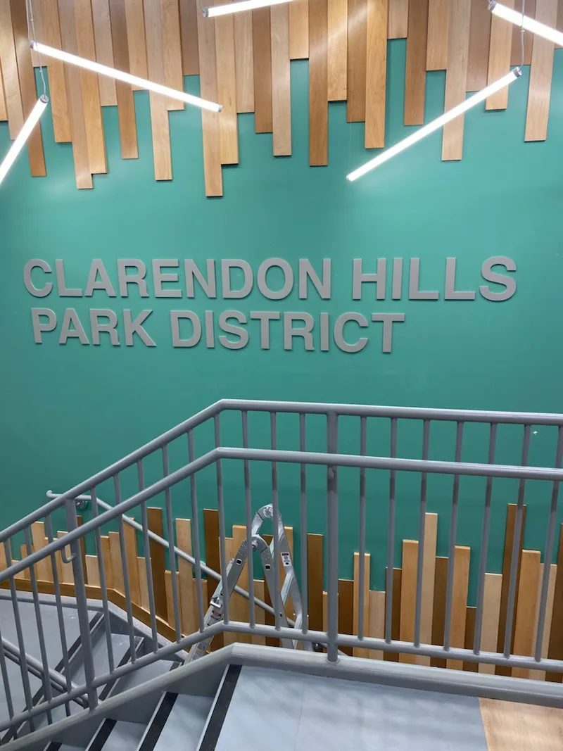 Clarendon Hills Park District interior — large dimensional metal letters across a teal-green stairwell wall framed by wood paneling and modern LED lighting.