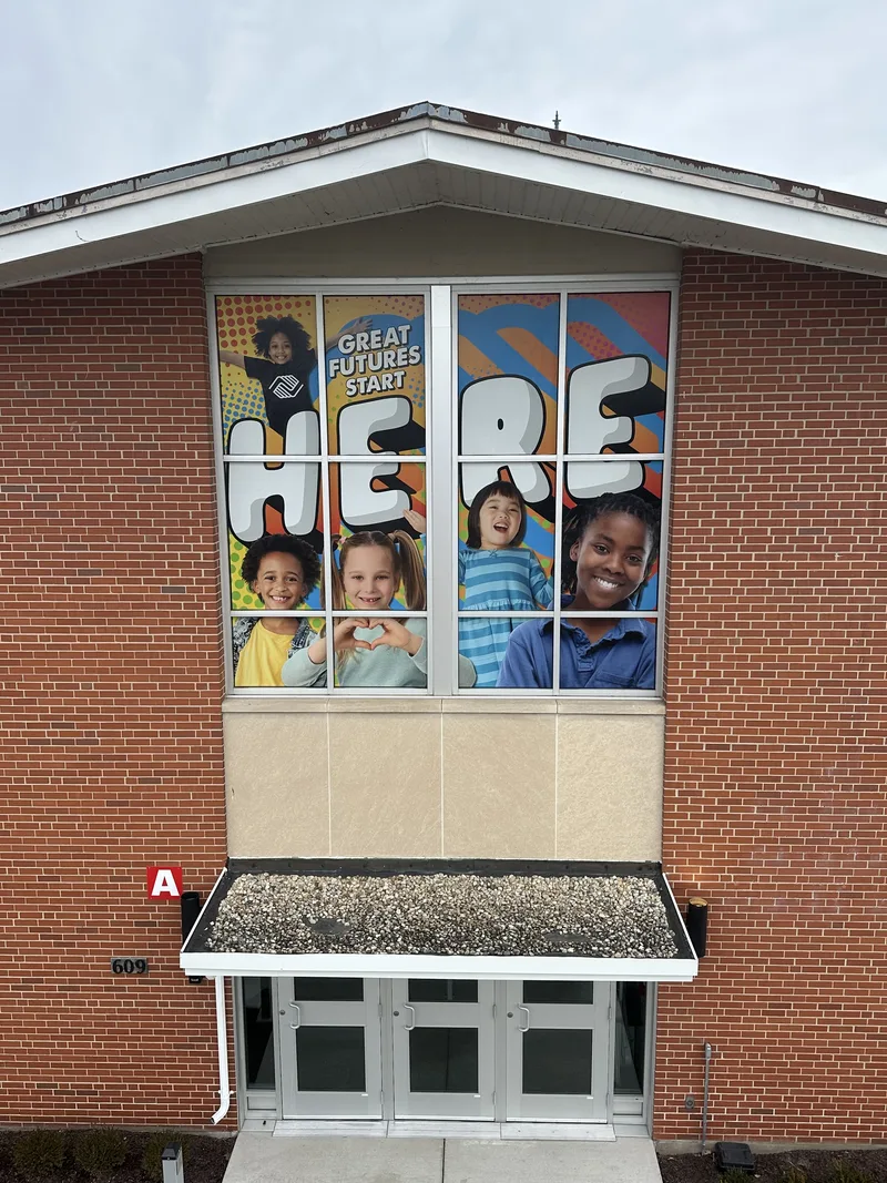Two-story window graphic — bold "HERE" lettering with smiling kids and "Great Futures Start Here" across a gridded window above a school-building entry.