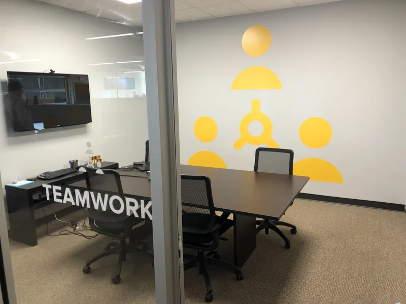 Comply365 Teamwork conference room — yellow people-and-target wall graphic with "TEAMWORK" frosted-glass door inscription.