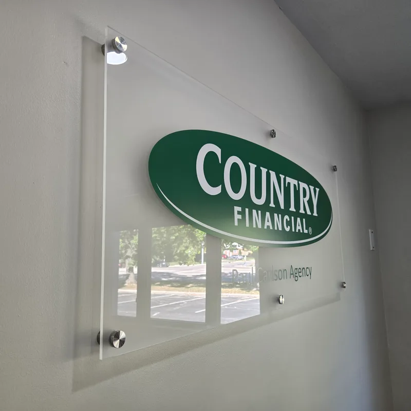 Country Financial (Mulford Rd agency) interior lobby sign — acrylic stand-off panel with green oval 'COUNTRY FINANCIAL' logo and 'Paul Carlson Agency' name on a frosted-edge clear acrylic, mounted with shiny chrome stand-offs against a white wall.