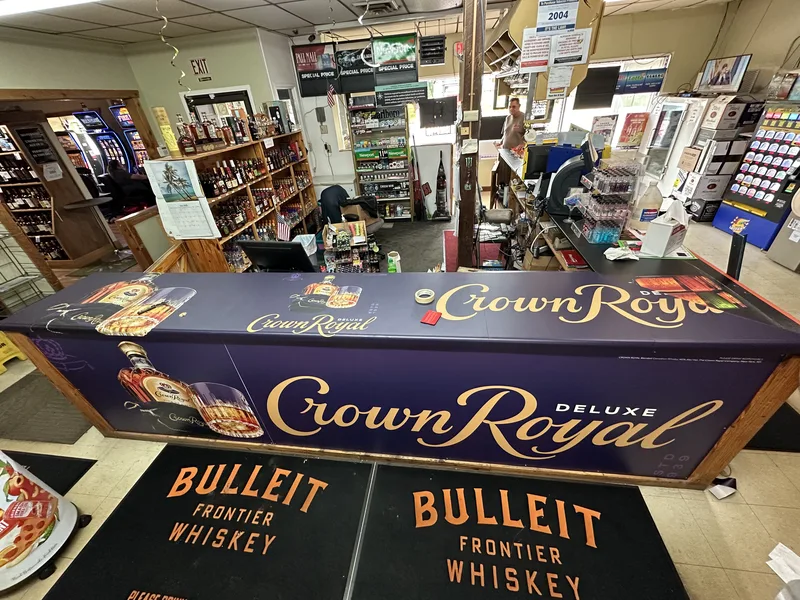 Liquor-store checkout counter wrapped in a full-color Crown Royal Deluxe graphic, with branded Bulleit floor mats in front.