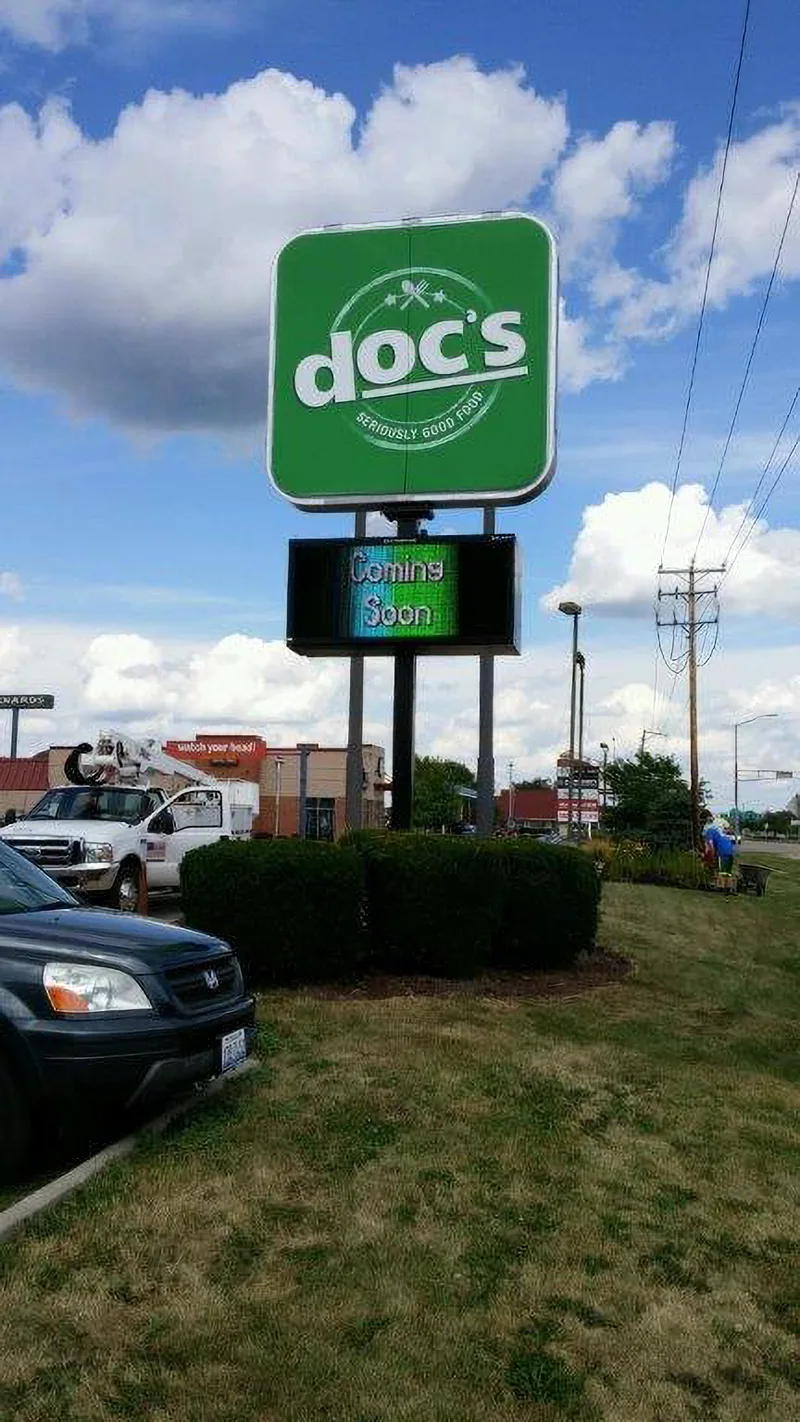 Doc's roadside pylon — green illuminated circular logo cabinet over a full-color LED message center, with the CorPro bucket truck in the background.