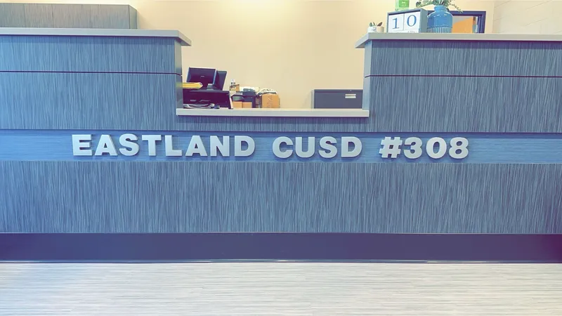 Eastland CUSD #308 reception desk — brushed-metal dimensional 'EASTLAND CUSD #308' letters mounted centered on a modern wood-grain reception counter, with an office phone and desk items visible behind.