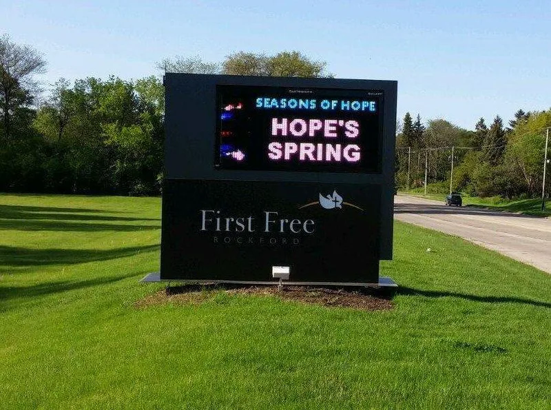 First Free Rockford church monument sign — modern black cabinet with dimensional 'First Free ROCKFORD' wordmark, dove-and-cross emblem, and full-color LED message center (showing 'SEASONS OF HOPE / HOPE'S SPRING'), set into a landscaped lawn on a sunny day.