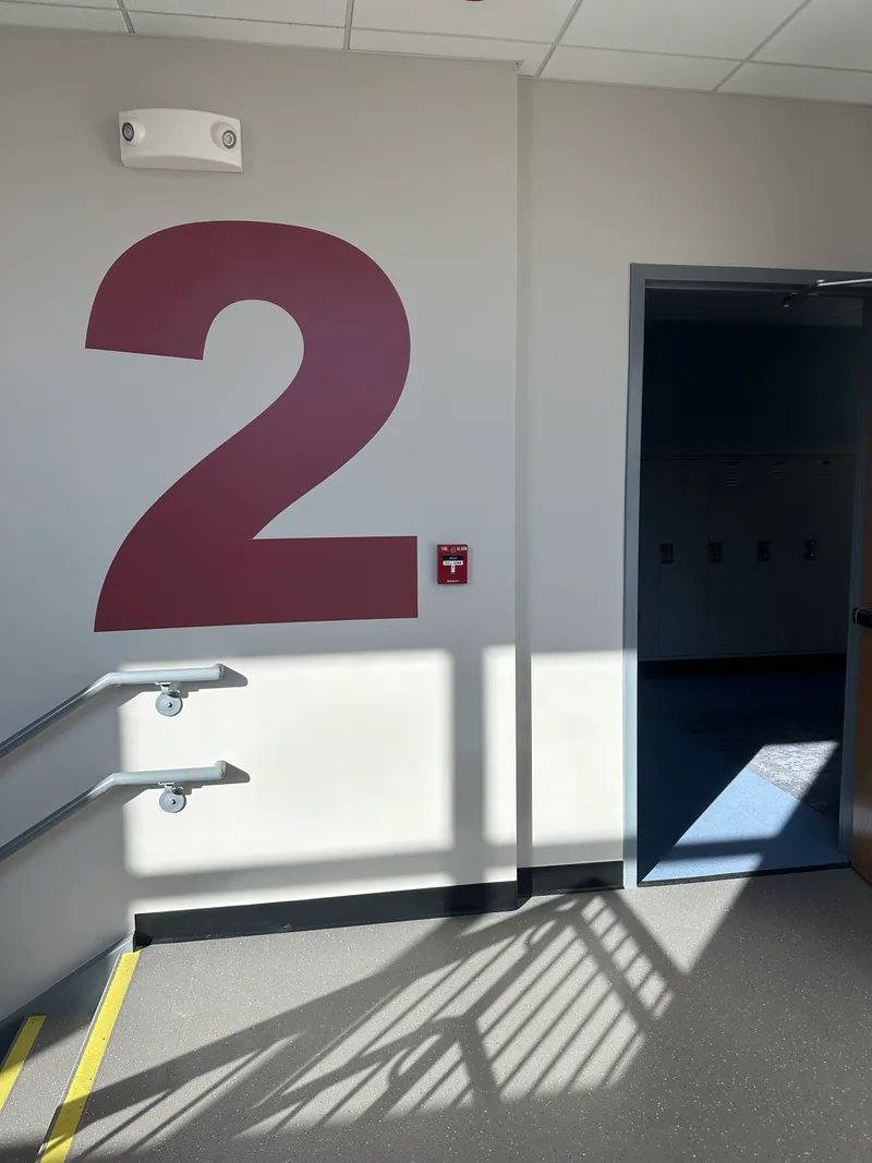 Modern school stairwell — oversized dimensional dark-red '2' floor identifier painted on a clean white wayfinding wall beside a doorway opening into a hallway of lockers.
