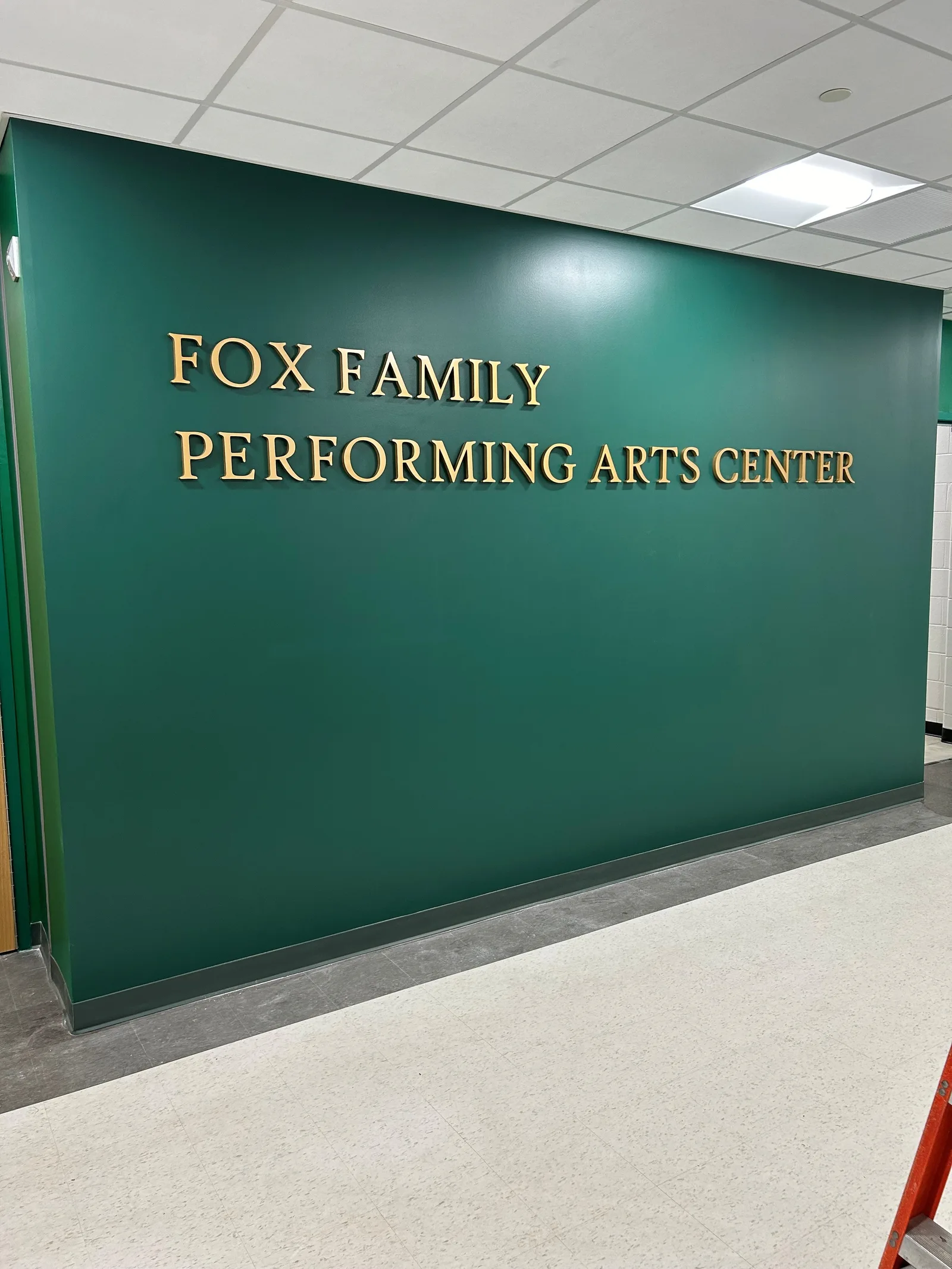 Fox Family Performing Arts Center — gold dimensional letters reading 'FOX FAMILY PERFORMING ARTS CENTER' mounted on a deep forest-green interior wall above a terrazzo floor.