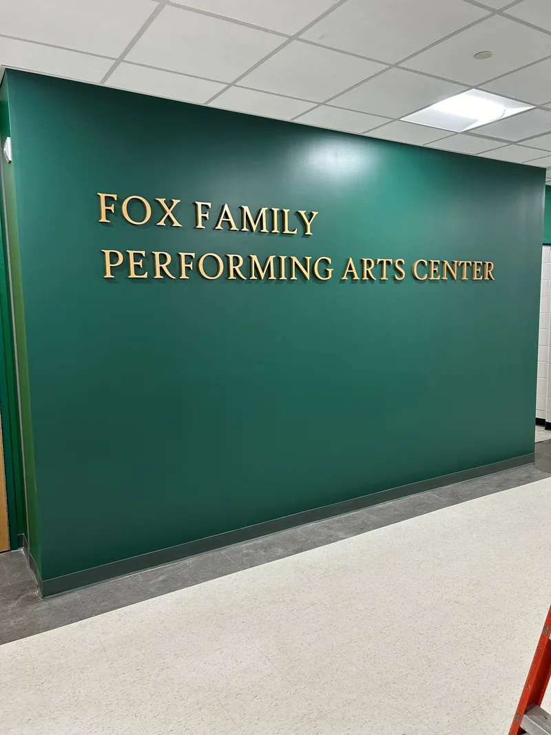 Fox Family Performing Arts Center — gold dimensional letters reading 'FOX FAMILY PERFORMING ARTS CENTER' mounted on a deep forest-green interior wall above a terrazzo floor.