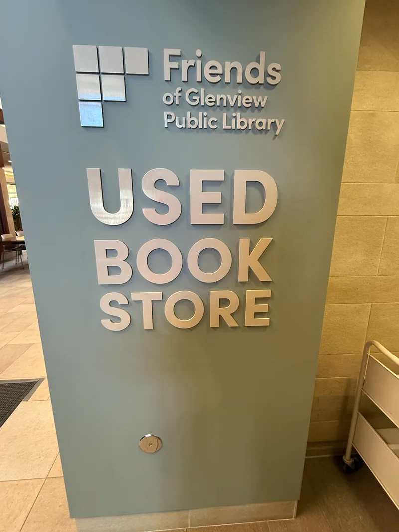 Glenview Public Library Used Book Store sign — sage-green dimensional pedestal panel with brushed-aluminum 'USED BOOK STORE' lettering and a Tetris-style cluster of square tiles forming the Friends of Glenview Public Library logo.