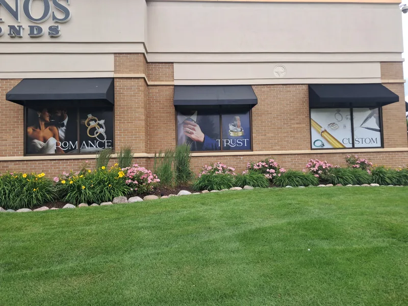 Gruno's Diamonds storefront window graphic install — three coordinated black-framed window panels ('Romance', 'Trust', 'Custom') featuring bride-and-groom, hand-with-wedding-bands, and custom-ring photography over a landscaped brick facade with dark awnings, blooming flowers, and manicured lawn.