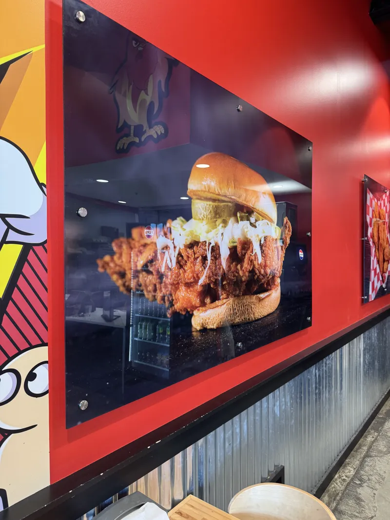 Hangry Joe's photorealistic food-graphic wall panel — large trapezoidal red-framed panel featuring an oversized printed image of a Hangry Joe's chicken sandwich, mounted on the corrugated-steel interior dining-room wall.