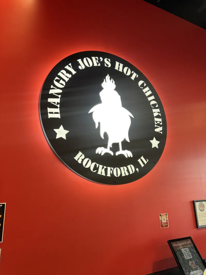 Hangry Joe's Rockford dimensional interior medallion — large circular black cutout logo with 'HANGRY JOE'S HOT CHICKEN / ROCKFORD, IL' arched around a bold rooster silhouette and star icons, mounted on a bright red interior wall.