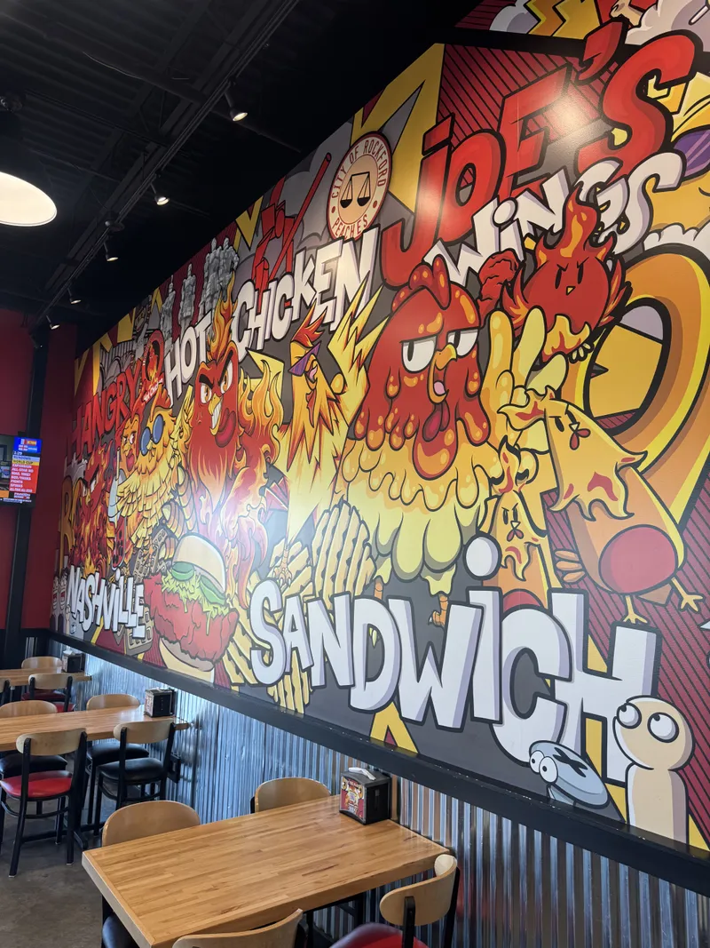 Hangry Joe's comic-book-style dining wall mural — vivid graffiti-inspired 'HANGRY / HOT CHICKEN / WINGS / JOE'S' typography surrounded by exaggerated rooster mascots breathing flames, City of Rockford Peaches seal, and 'NASHVILLE / SANDWICH' callouts, printed at full room height across the dining room.