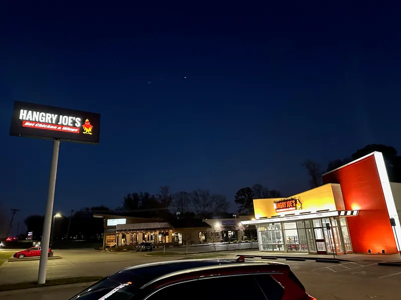 Hangry Joe's Hot Chicken & Wings storefront at night — dramatic dusk shot of the Tyler, TX location with an illuminated pylon sign displaying the red-and-white 'Hangry Joe's' rooster logo in the foreground, and the warm-lit orange-red restaurant storefront in the background with matching illuminated cabinet signage.