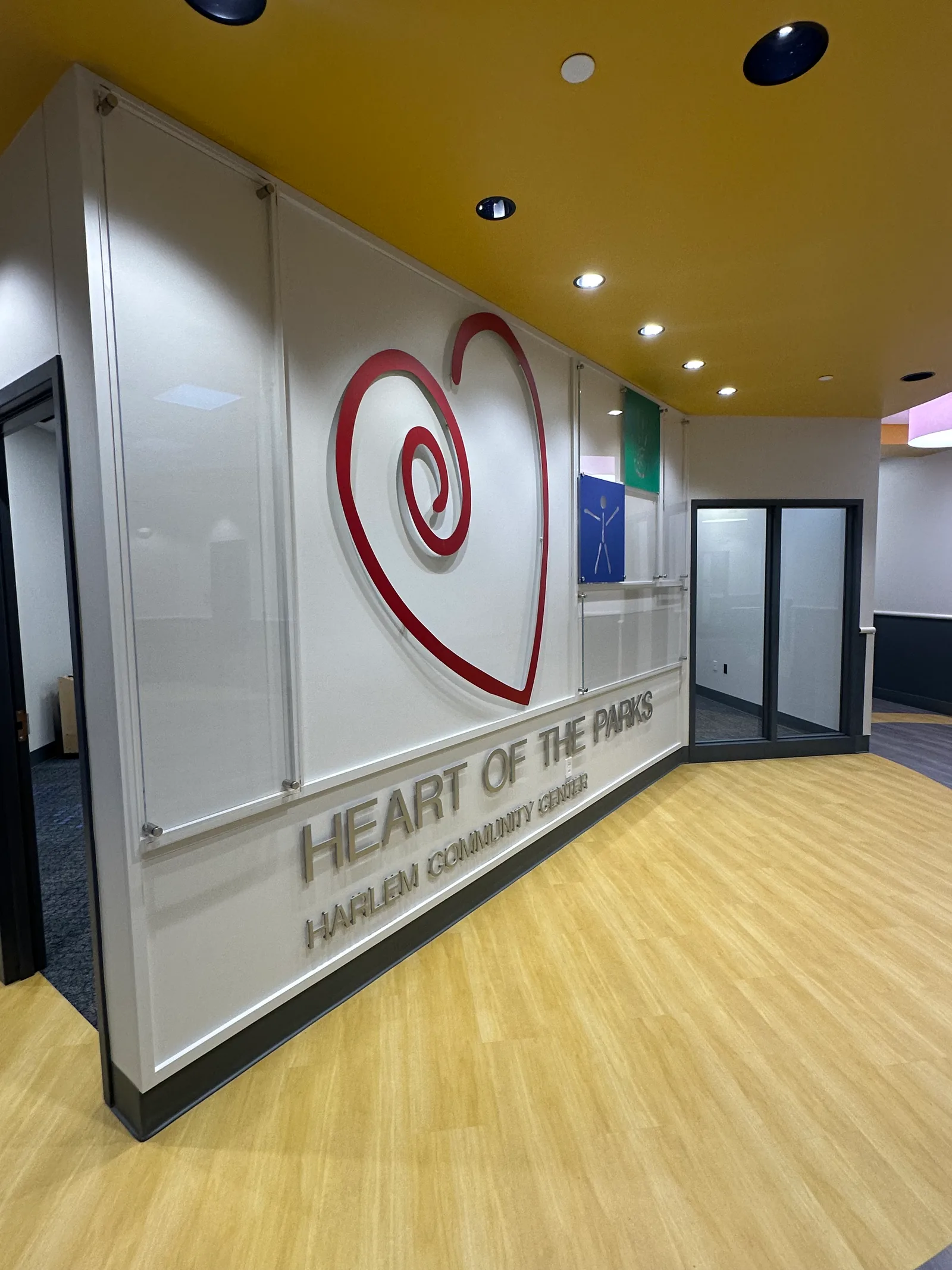 Harlem Community Center entry — acrylic stand-off 'Heart of the Parks — Harlem Community Center' dimensional sign with a red swirling-heart logo mounted on a clean white frame, set into a warm yellow accent wall and wood-paneled wall.
