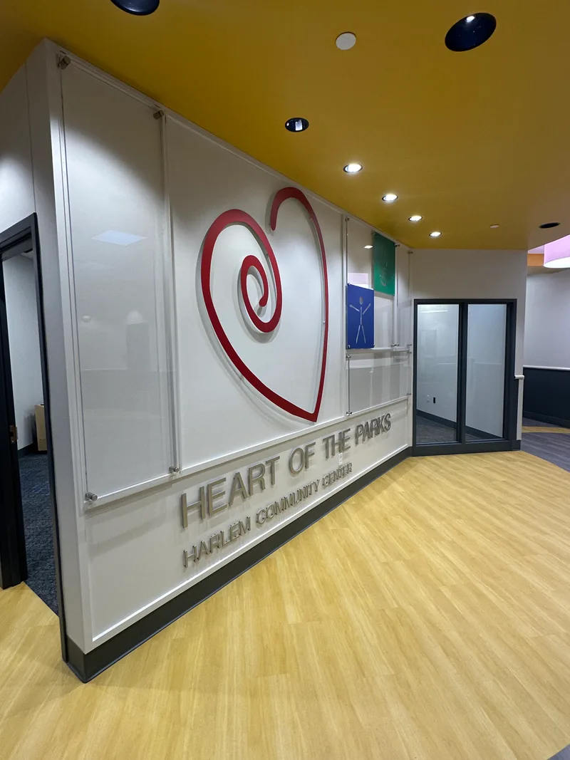 Harlem Community Center entry — acrylic stand-off 'Heart of the Parks — Harlem Community Center' dimensional sign with a red swirling-heart logo mounted on a clean white frame, set into a warm yellow accent wall and wood-paneled wall.