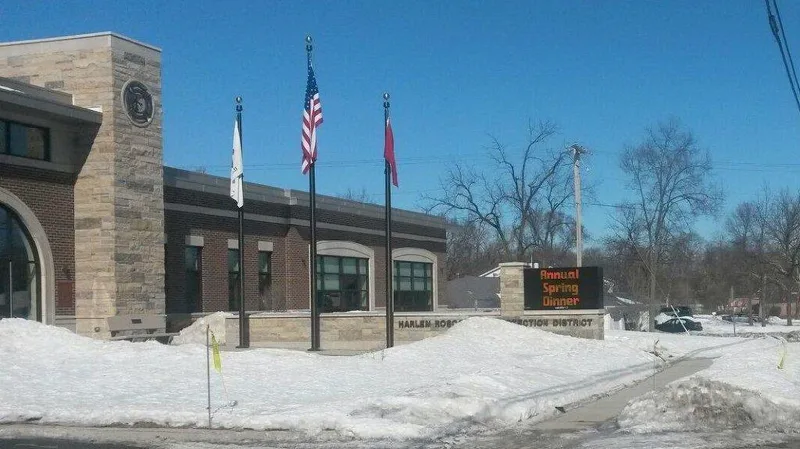 Harlem-Roscoe Fire Protection District station — brick exterior with carved-stone seal, flagpoles, and an LED community-message monument out front.