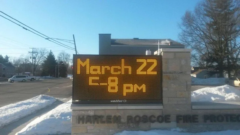 Harlem-Roscoe Fire Protection District stone-clad monument with a large amber LED message center reading "March 22 5-8 pm", set in winter light.