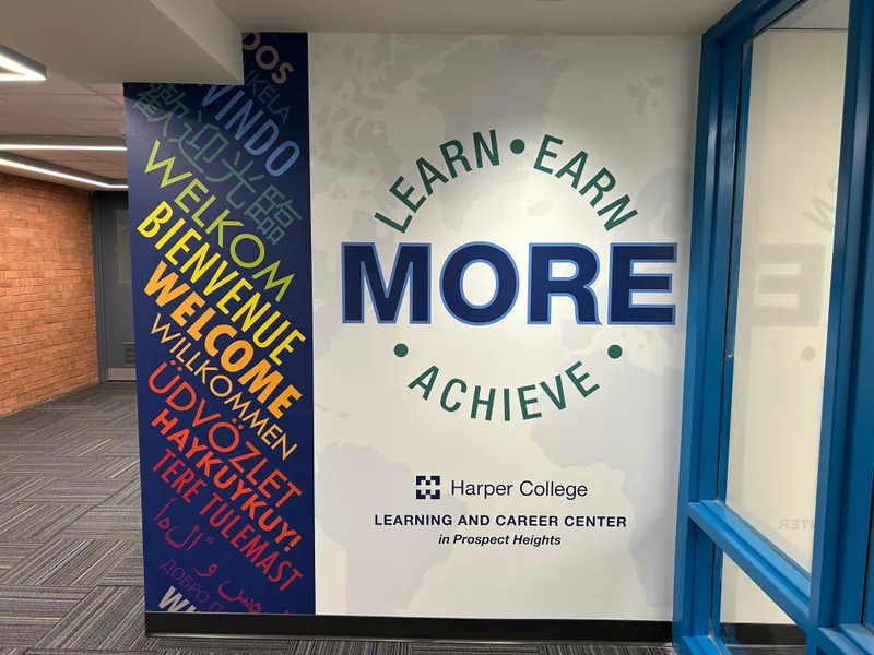 Harper College Learning and Career Center wall graphic (Prospect Heights) — 'Learn · Earn · MORE · Achieve' over a soft world-map background, paired with a full-height navy column of 'Welcome' translated into a dozen languages in vibrant colors.