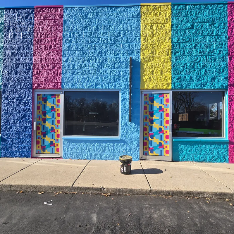 HCCAC Hazel Crest storefront — bold vertically-striped painted-brick facade in magenta, blue, yellow, and cyan with matching geometric CMYK-patterned window graphics on the storefront doors and side lights.