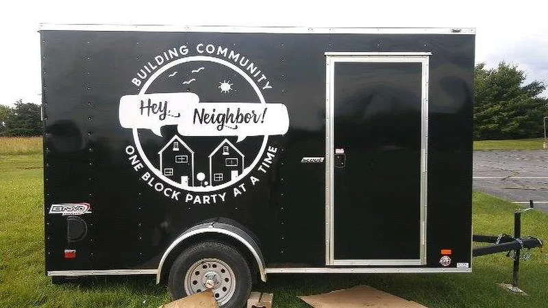 Black enclosed cargo trailer with a white circular "Building Community — Hey, Neighbor! — One Block Party at a Time" vinyl graphic.