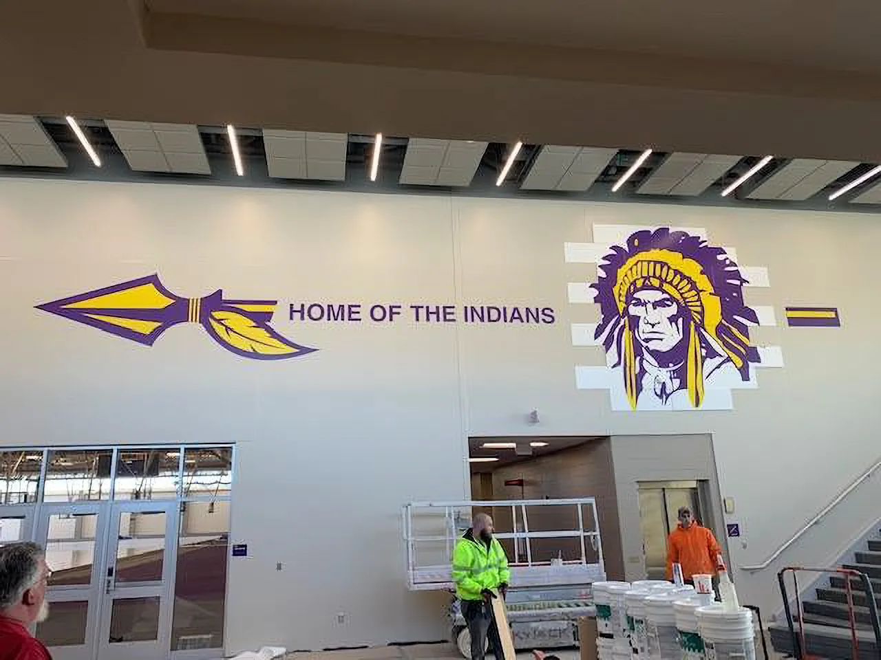 Hononegah High School field house — purple-and-gold "HOME OF THE INDIANS" spear graphic and mascot logo on a tall white wall, with the install crew and lift below.