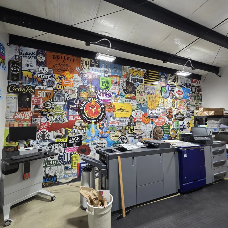 Interstate Graphics production office — floor-to-ceiling collage wall graphic of hundreds of client logos and labels behind the digital press line.