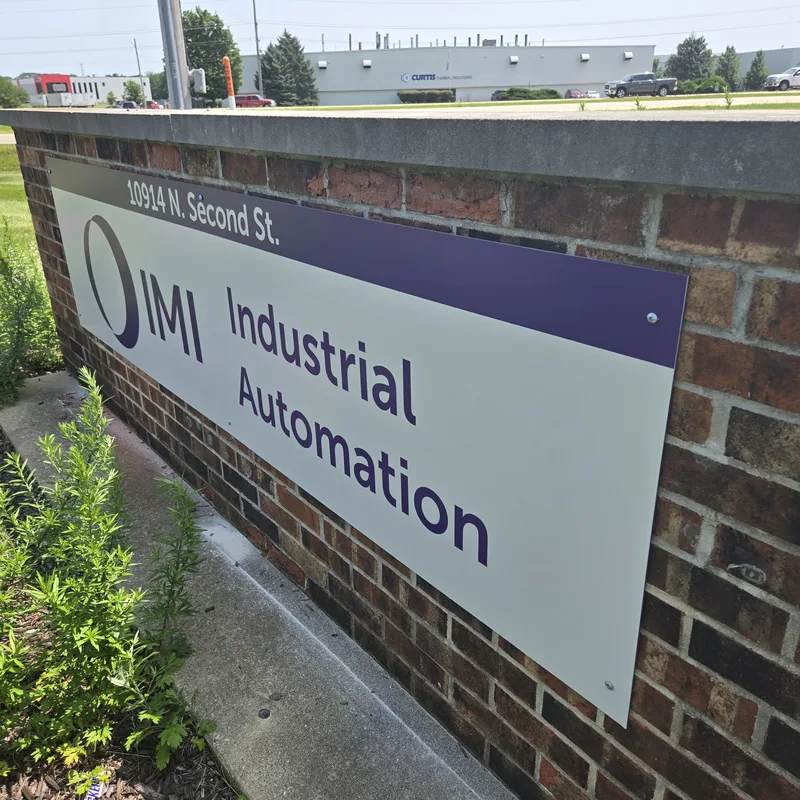 IMI Industrial Automation address panel — white aluminum face with purple header, "10914 N. Second St." and the IMI ring logo, mounted to a brick monument wall.