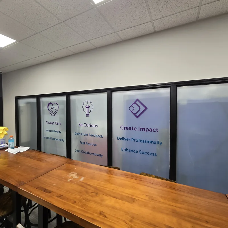 IMI break-room glass wall — frosted window film with purple icons and core values ("Always Care", "Be Curious", "Create Impact") above a long wood counter.