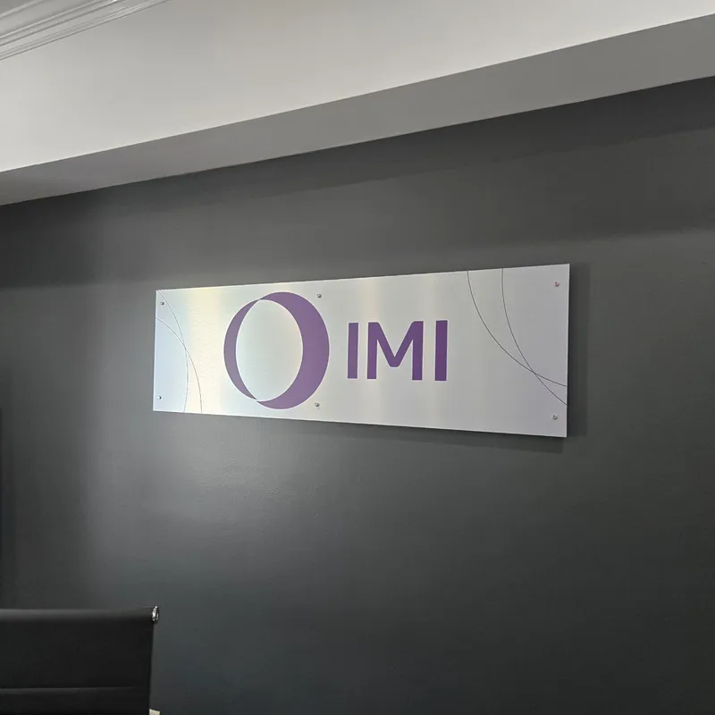 IMI interior wall sign — brushed-metal panel with cutout 'O IMI' purple mark (circle with crescent) and subtle etched line detail, mounted flush on a modern dark-gray office wall.