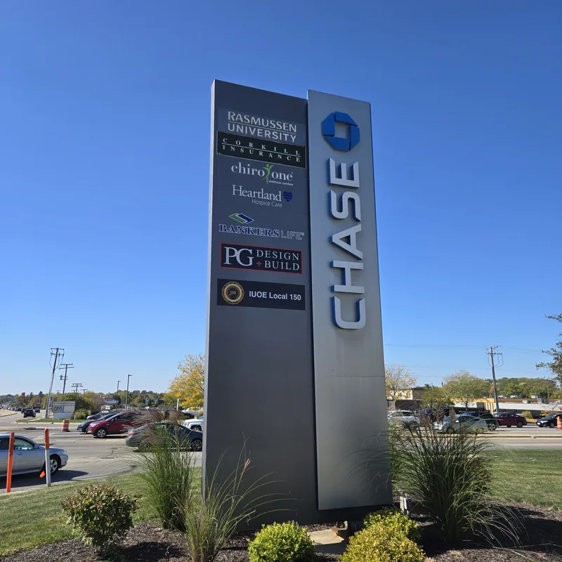 Tall aluminum multi-tenant pylon with a vertical blue "CHASE" blade and tenant panels for Rasmussen University, Chiro One, Heartland Dental, PG Design + Build and IUOE Local 150, in a landscaped island under blue sky.