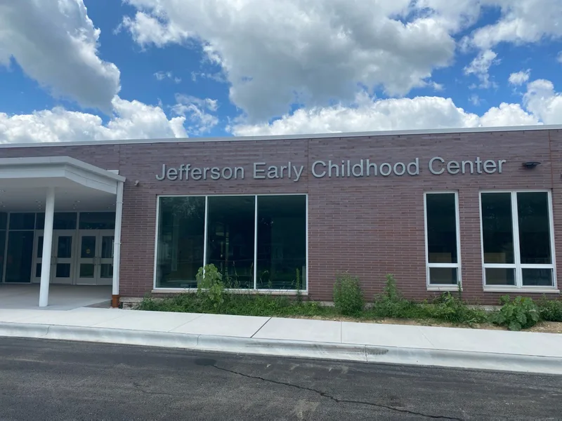 Jefferson Early Childhood Center — brushed-aluminum dimensional letters across a red-brick facade above the entry canopy under a bright cloudy sky.