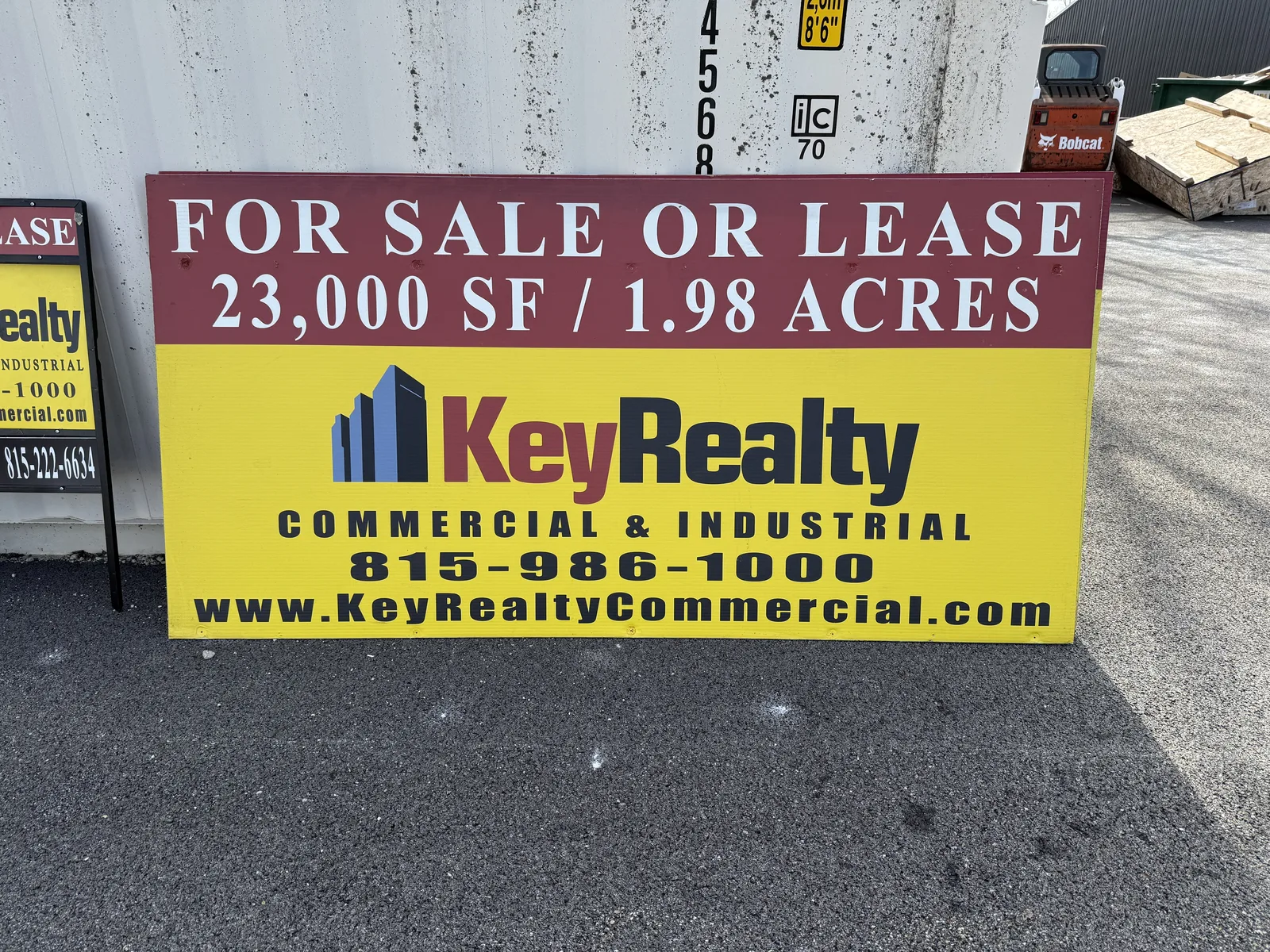 Key Realty commercial real-estate sign — "FOR SALE OR LEASE 23,000 SF / 1.98 ACRES" header on a bold yellow panel with the Key Realty logo, phone and web address.