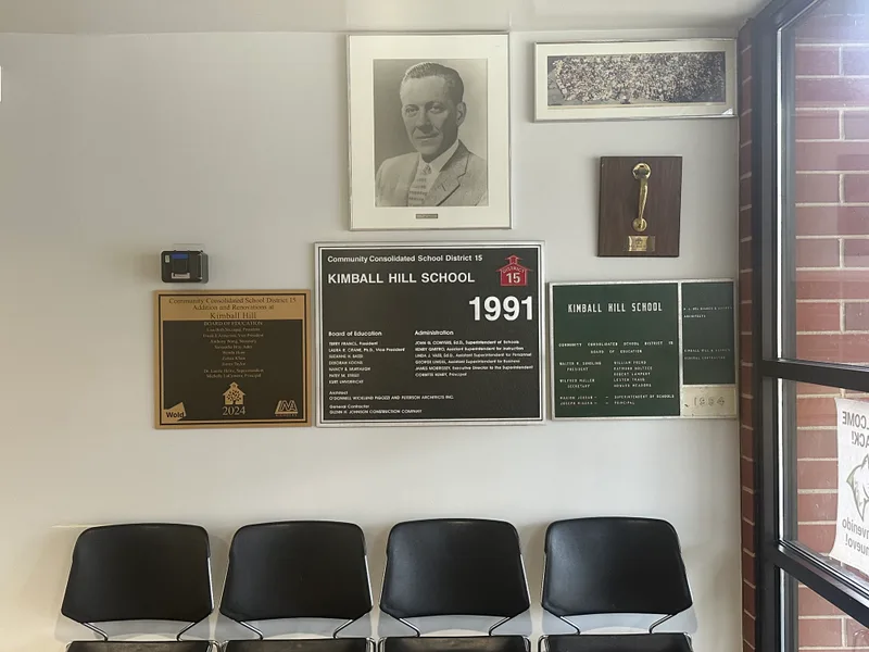 Kimball Hill School (Community Consolidated School District 15) lobby plaque wall — collection of dedication plaques spanning 1954 to 2024 (board members, principals, architects, contractors) arranged beneath a black-and-white portrait of the building's namesake.