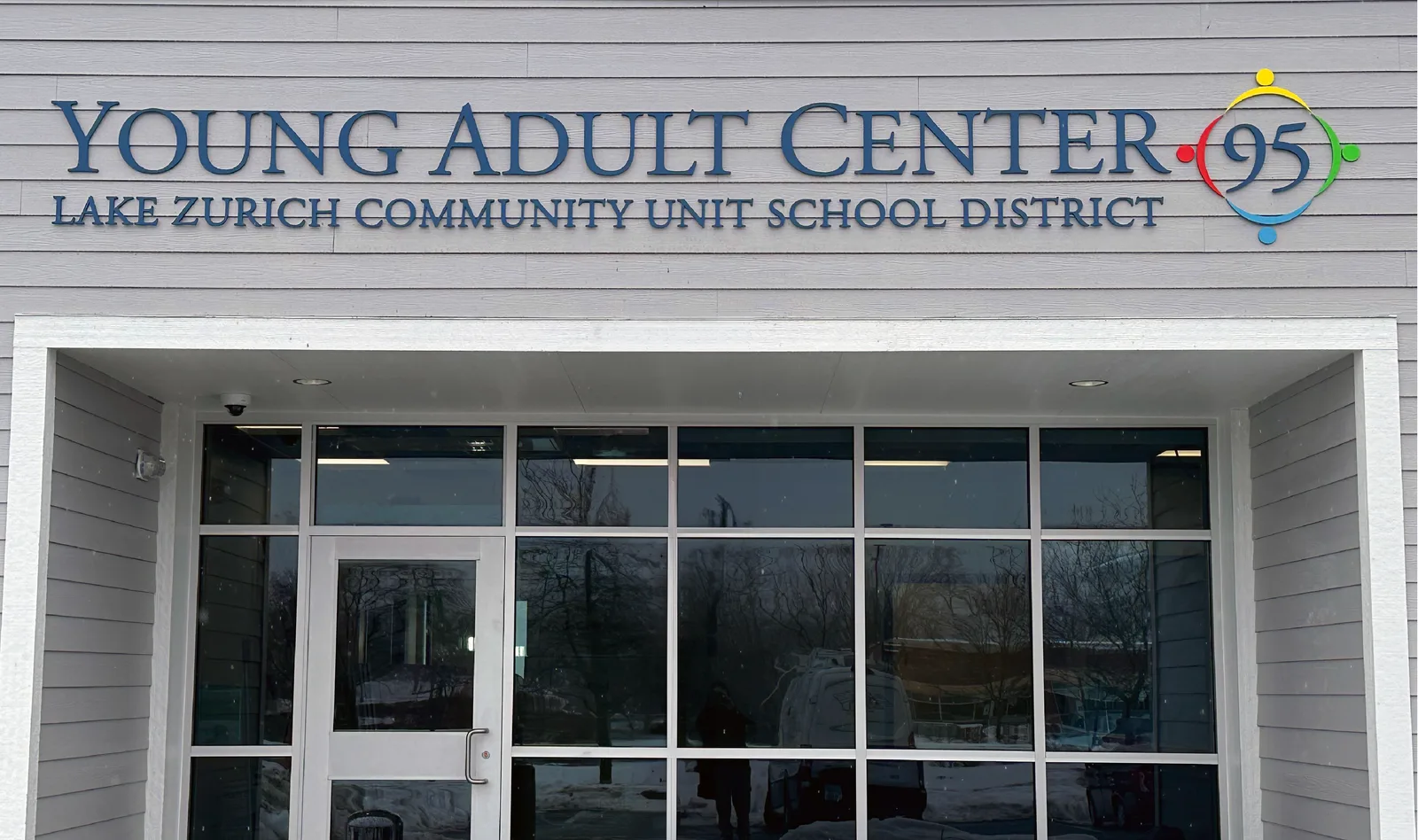 Lake Zurich Community Unit School District 95 Young Adult Center — clean exterior dimensional letters 'YOUNG ADULT CENTER · 95' with 'LAKE ZURICH COMMUNITY UNIT SCHOOL DISTRICT' subline, paired with a colorful circular logo of figures joining hands, mounted on the gray clapboard facade above the glass entry.