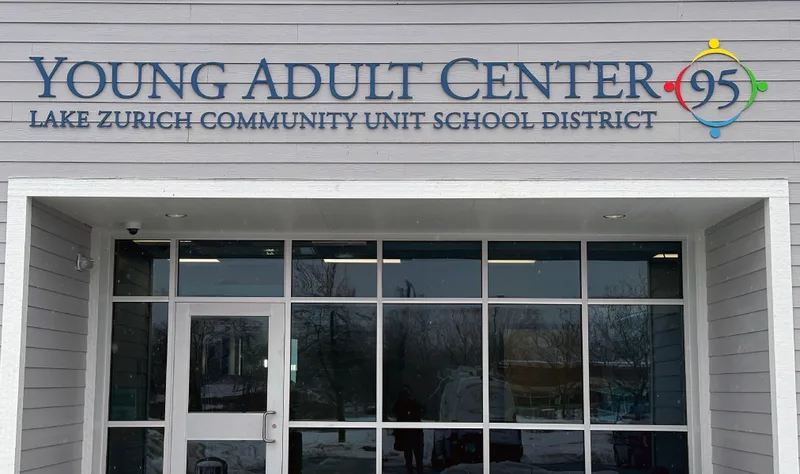 Lake Zurich Community Unit School District 95 Young Adult Center — clean exterior dimensional letters 'YOUNG ADULT CENTER · 95' with 'LAKE ZURICH COMMUNITY UNIT SCHOOL DISTRICT' subline, paired with a colorful circular logo of figures joining hands, mounted on the gray clapboard facade above the glass entry.