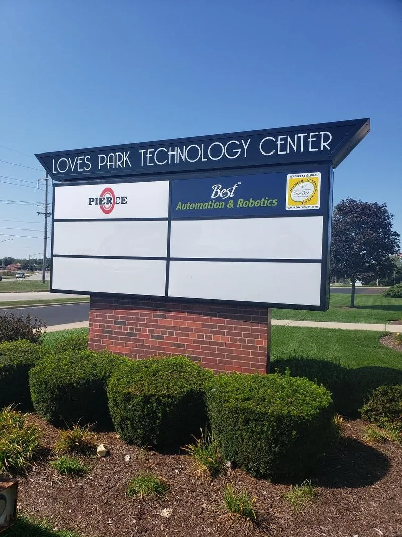 Loves Park Technology Center multi-tenant monument sign — angled navy cabinet with 'LOVES PARK TECHNOLOGY CENTER' header and replaceable tenant panels (Pierce, Best Automation & Robotics, TeamBest Global) on a brick base surrounded by landscape shrubs.