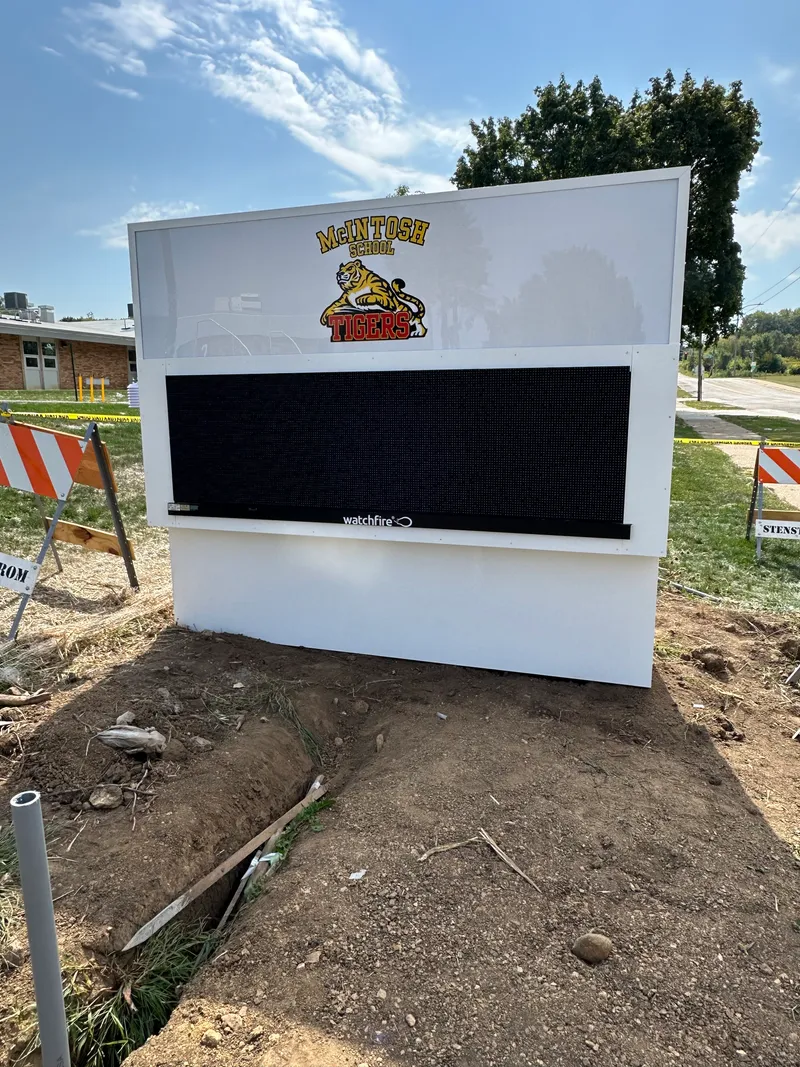 McIntosh School monument — freshly installed roadside monument with dimensional gold-and-black 'McIntosh School Tigers' tiger-mascot cabinet on a white base, Watchfire LED message center integrated below, mounted on a concrete plinth with fresh soil and construction barricades still in view.