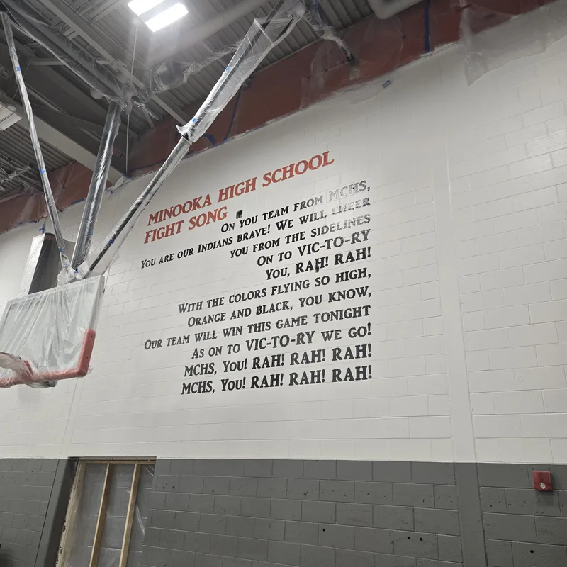 Minooka Community High School gym — the full school fight song applied in red and black lettering across the white block wall above the baseline.